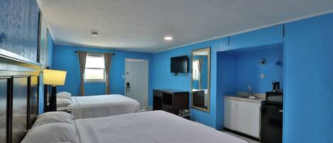 A hotel room with two beds, a desk with a lamp, a small kitchenette, and a mirror on the wall.
