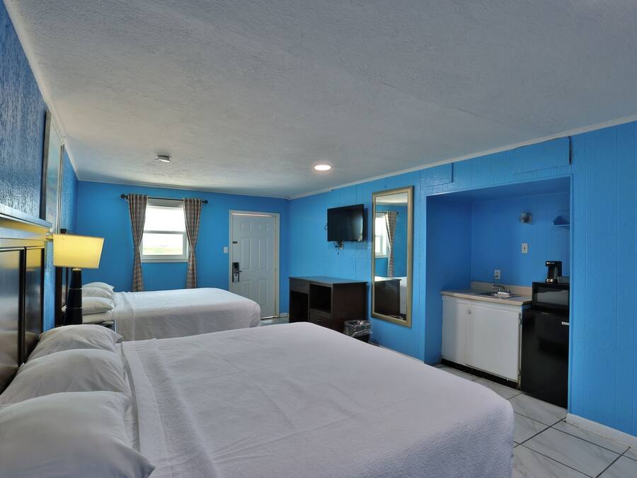 A hotel room with two beds, a desk with a lamp, a small kitchenette, and a mirror on the wall.