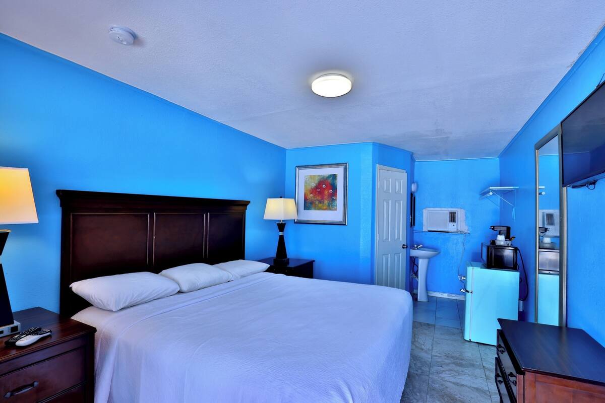 Habitación de hotel con cama, mesitas de noche, lámpara, un cuadro, lavabo, refrigerador y espejo.