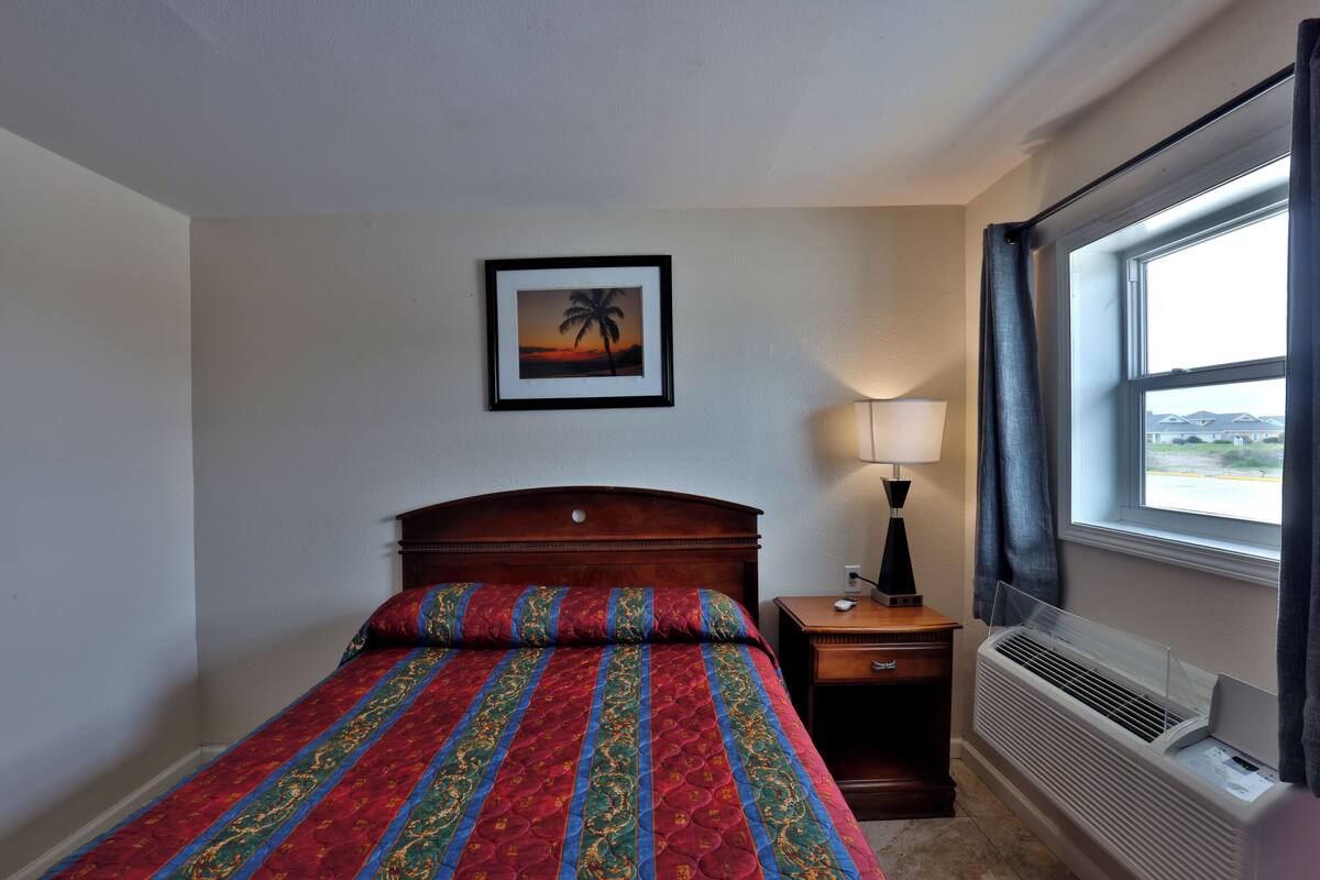 Una habitación de hotel con cama, mesita de noche, lámpara, ventana con cortinas e imagen colgada en la pared.