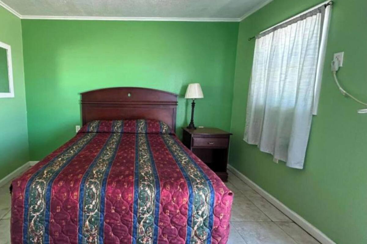 Un dormitorio con cama, mesita de noche, lámpara y ventana con cortinas.