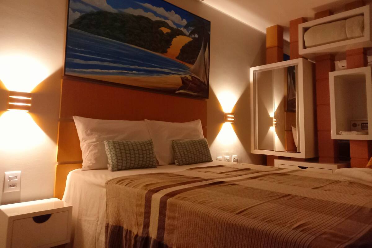 Habitación de hotel con cama, almohadas, un cuadro en la pared y luces empotradas.