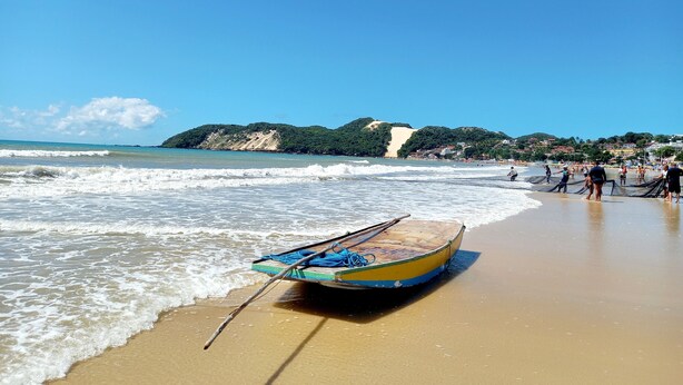 A beach with a rowboat on the sand, waves gently lapping the shore, and a distant view of a hilly landscape with buildings. - Ilusion Flats Aconchego de Ponta Negra (Natal)