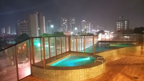 A rooftop pool area with tiled flooring and glass railings, surrounded by city buildings at night. - Ilusion Flats Aconchego de Ponta Negra (Natal)
