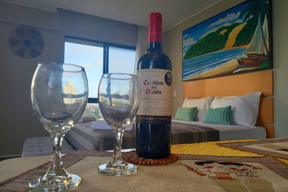 Una botella de vino Casillero del Diablo, dos copas y una mesa con un mantel individual en una habitación de hotel con una cama y un cuadro en la pared.