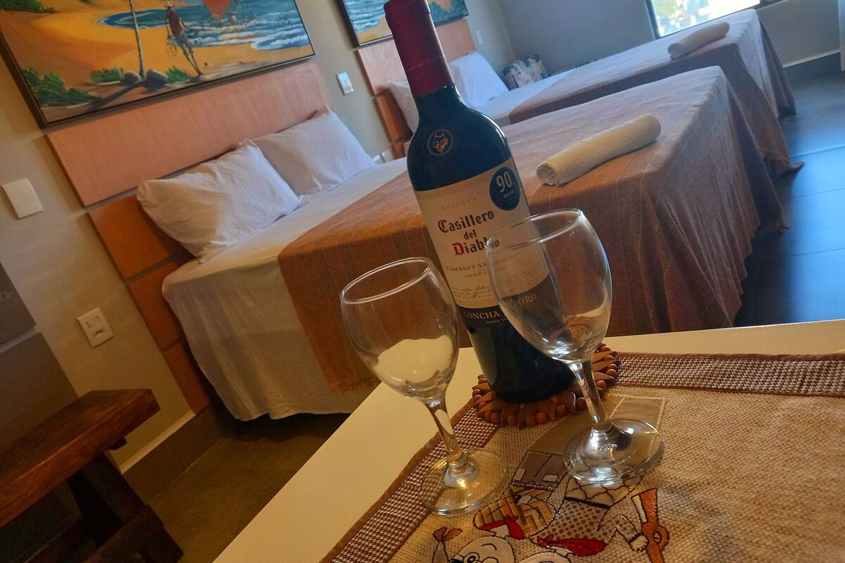 Habitación de hotel con cama, una mesa con una botella de Casillero del Diablo y dos copas, y vista al océano desde la ventana.