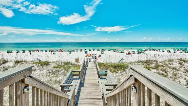 Een houten looppad dat naar een strand leidt, met parasols en mensen op het strand.