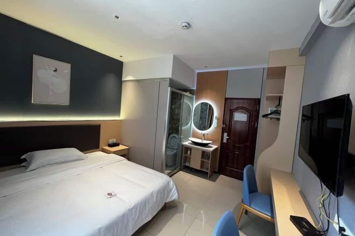 A hotel room with a bed, a desk with a chair, a TV, a mirror, and a bathroom with a sink and toilet.