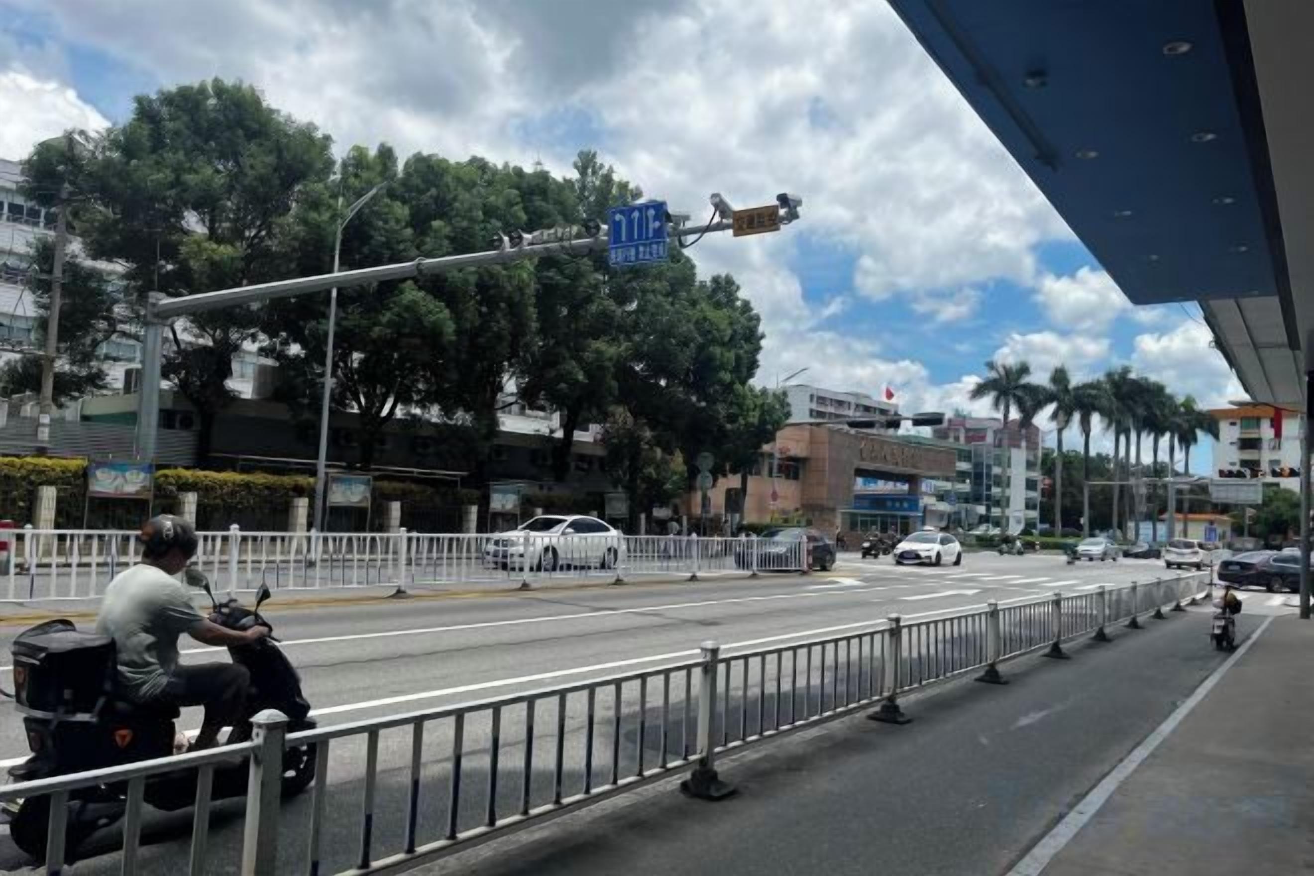 A street scene with a metal barrier, vehicles, and buildings in the background.