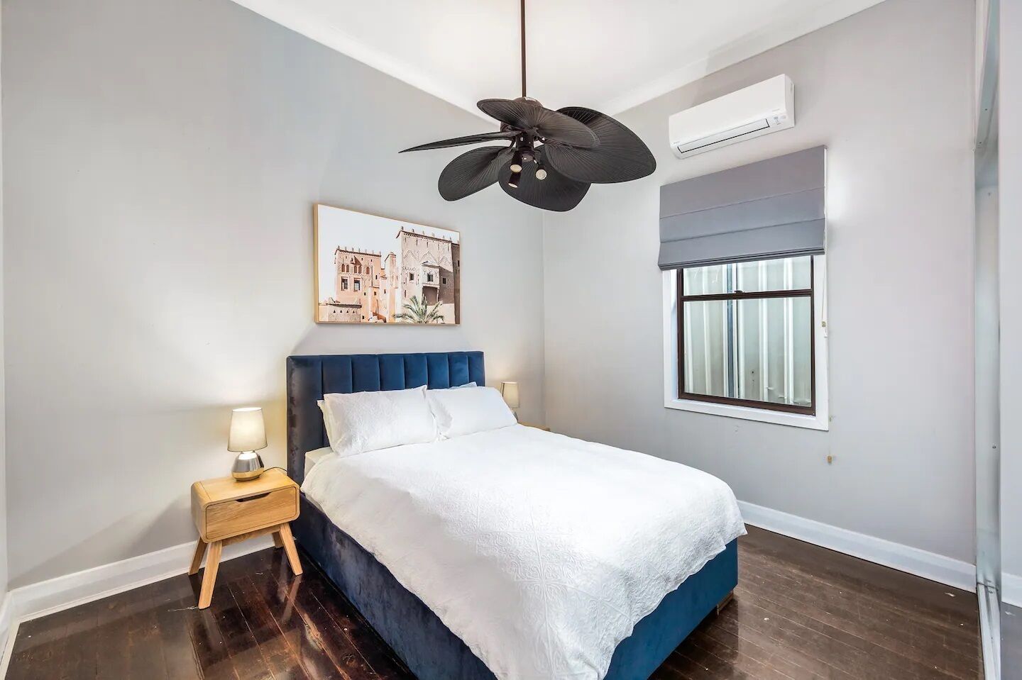 A bedroom with a bed, a nightstand, a ceiling fan, an air conditioning unit, a window, and a wall-mounted picture.