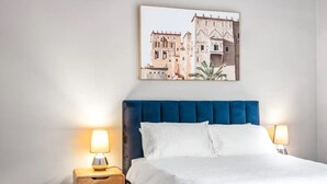 Een slaapkamer met een bekleed hoofdeinde in blauw, witte beddengoed en twee bedlampjes.