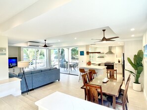 Amplia sala de estar con sofá, mesa de comedor y sillas. Hay una zona de cocina con refrigerador y ventilador de techo.