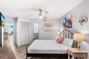 A bedroom with a bed, a ceiling fan, a nightstand, a lamp, a clock, and artwork on the wall. - Sandcastle Condominiums Unit 104 (Port Aransas)
