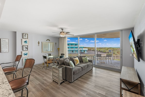 A modern living room with a gray sofa, wooden dining table, and chairs. A large window offers a view of a blue building and a clear sky. - Sandcastle Condominiums Unit 104 (Port Aransas)