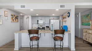 A modern kitchen with white cabinets, a central island with bar stools, a microwave, oven, and a refrigerator. - Sandcastle Condominiums Unit 104 (Port Aransas)