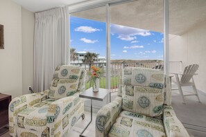 A balcony with two chairs, a small table with a vase, and a view of a beach and palm trees. - Sandcastle Condominiums Unit 106 (Port Aransas)