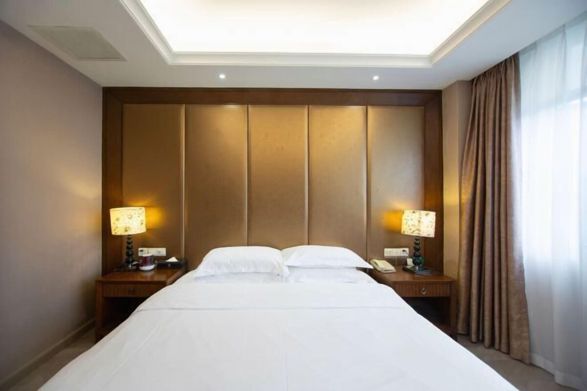 Habitación de hotel con una cama grande, dos mesitas de noche con lámparas, un cabecero y una ventana con cortinas.
