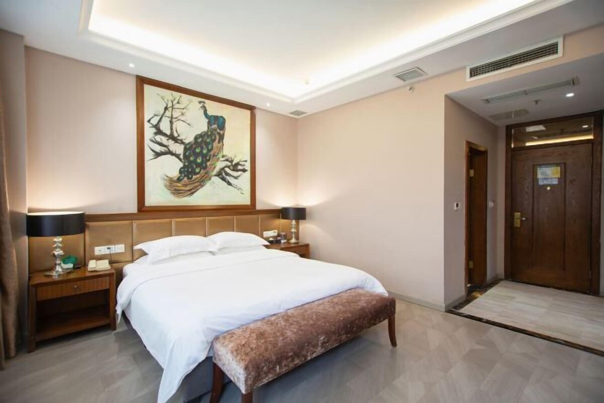 Habitación de hotel con una cama grande, mesitas de noche, un banco, un cuadro en la pared y una puerta de madera.