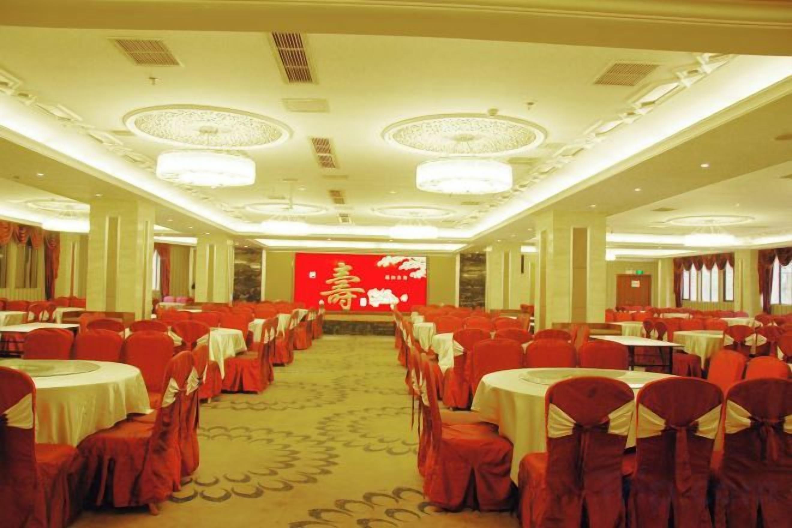 Un salón de banquetes con mesas y sillas redondas, un escenario con fondo rojo y suelo alfombrado.