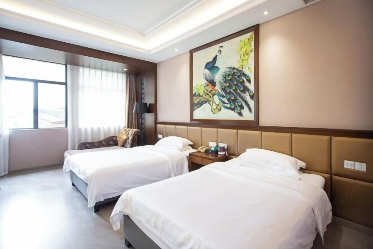 Habitación de hotel con dos camas, un cuadro de un pavo real, una ventana con cortinas y una mesita de noche con lámpara.