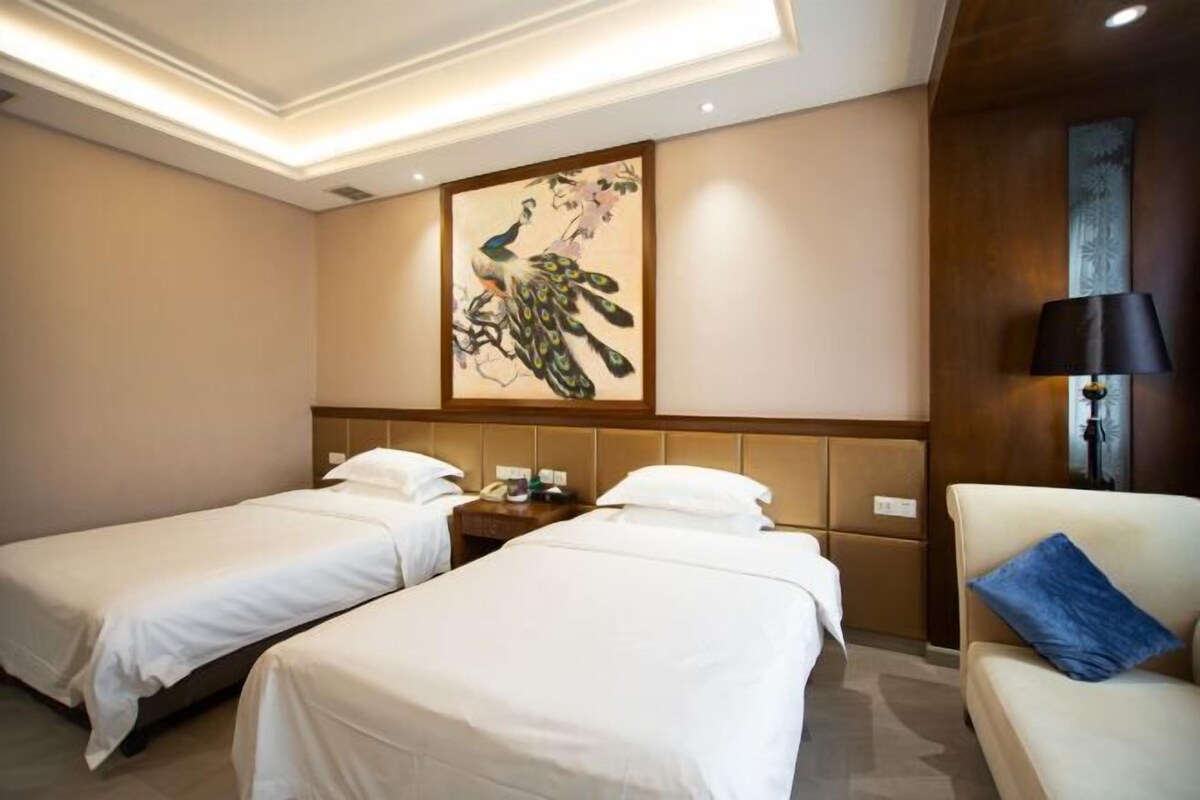 Habitación de hotel con dos camas individuales, cabecera de madera, un cuadro colgado en la pared, una mesita de noche con lámpara y un sofá con un cojín azul.