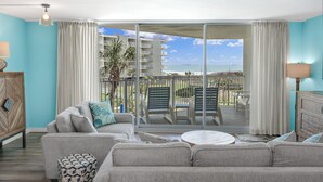 Una sala de estar con un amplio ventanal corredera que da a un balcón con vista a la playa y un edificio.