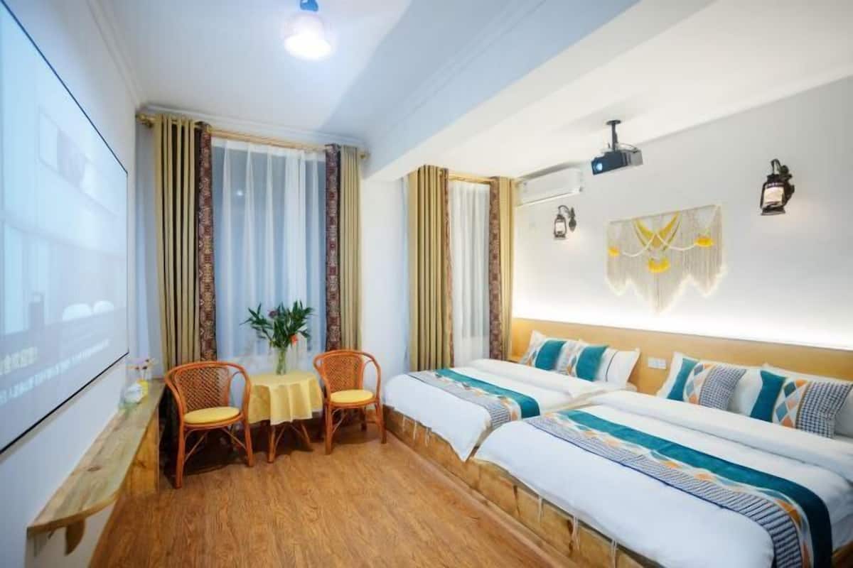A hotel room with two beds, a small table with chairs, a projector screen, and a wall-mounted air conditioner.