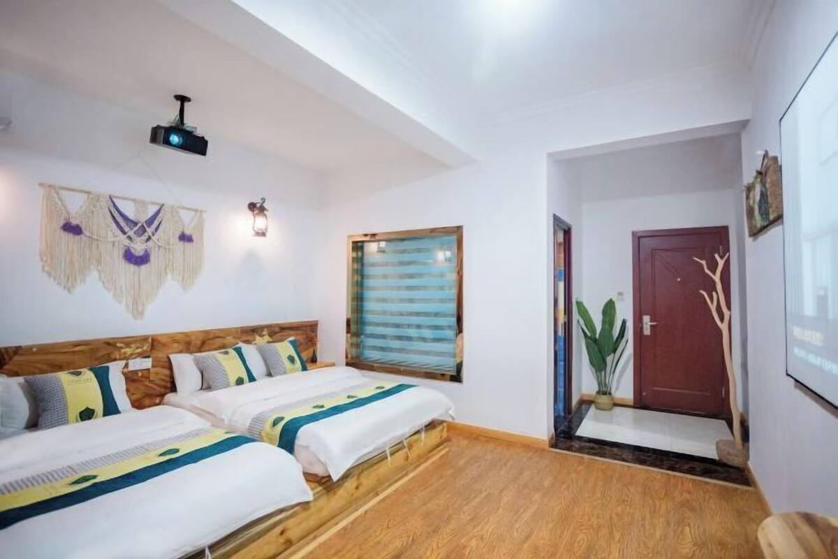A room with two beds, a wooden floor, a ceiling projector, a wall-mounted light, a window with blinds, and a door with a plant beside it.
