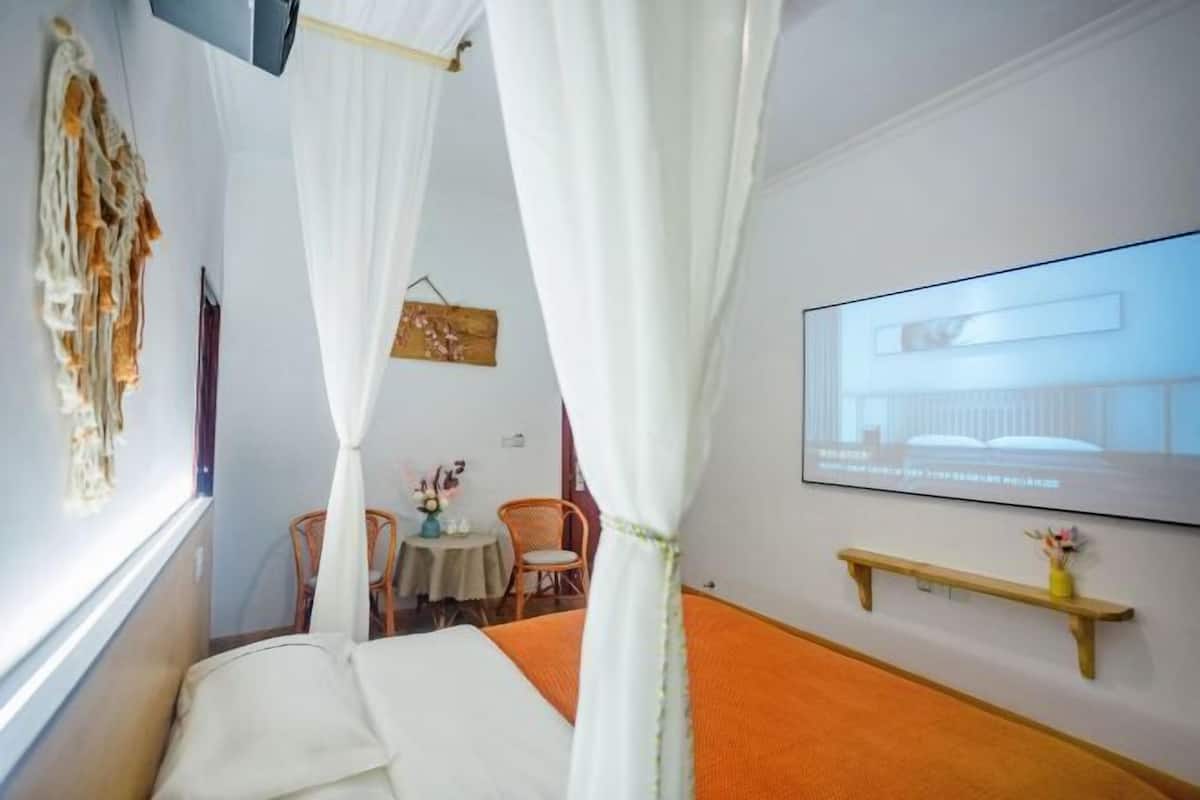A bedroom with a bed, a TV on the wall, a small table with chairs, and a window with curtains.