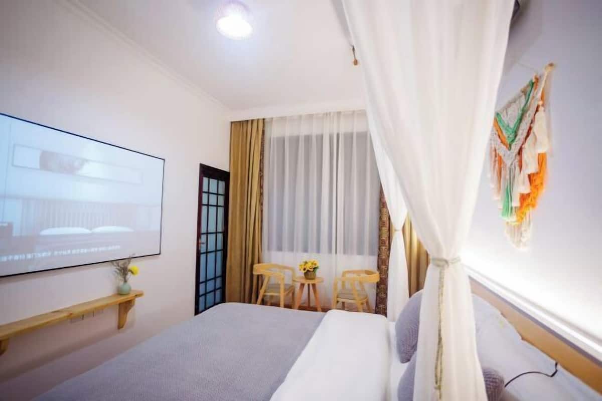 A hotel room with a bed, a TV on the wall, a small table with chairs, and a window with curtains.