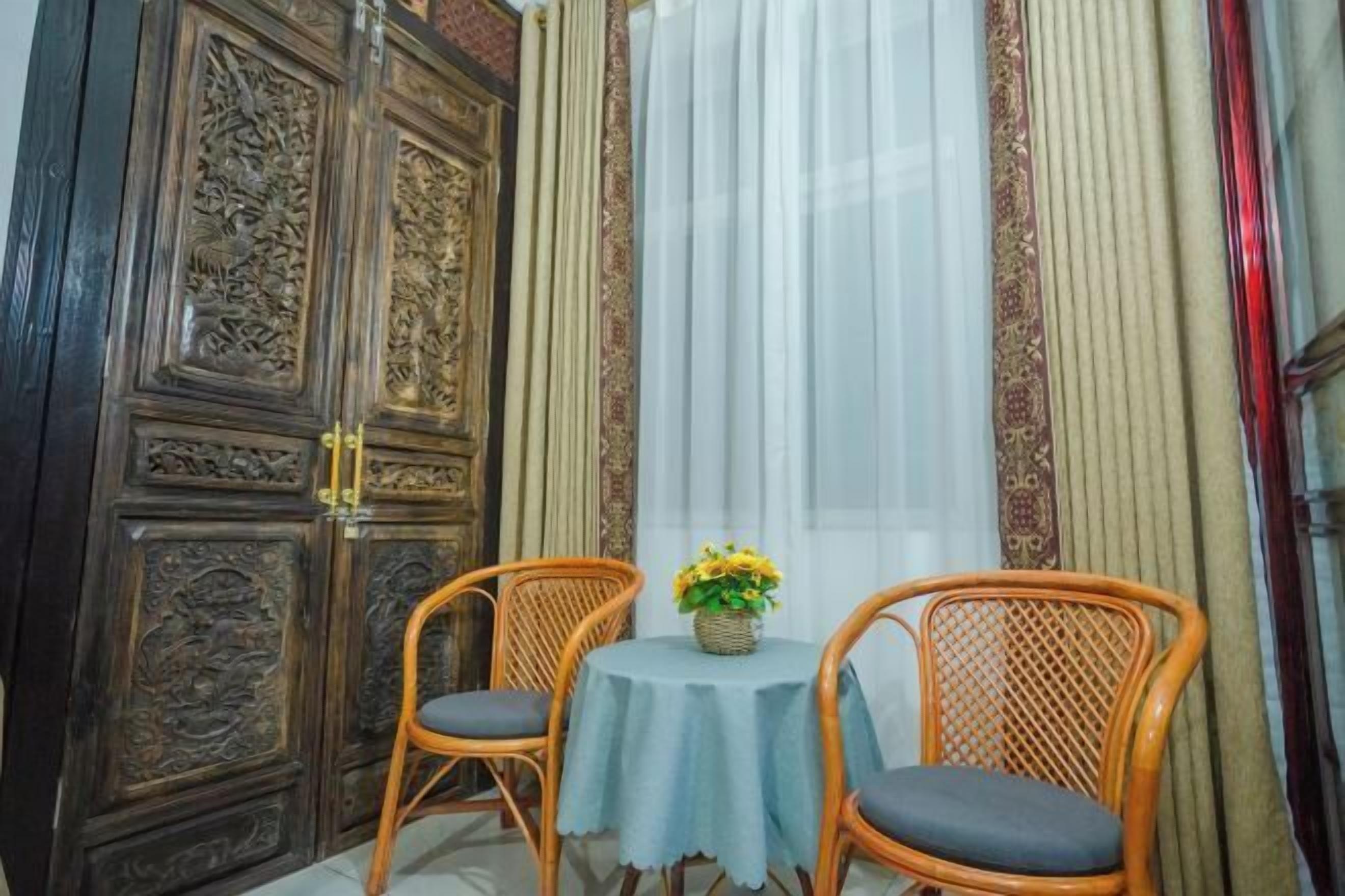 Una habitación con puerta de madera, dos sillas de mimbre, una mesita con mantel azul y una ventana con cortinas translúcidas.