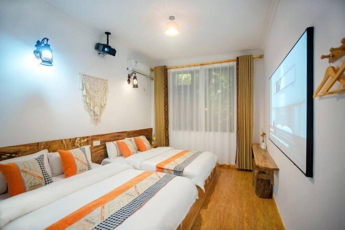 A hotel room with two beds, a wooden headboard, a wall-mounted projector, and a large window with curtains.