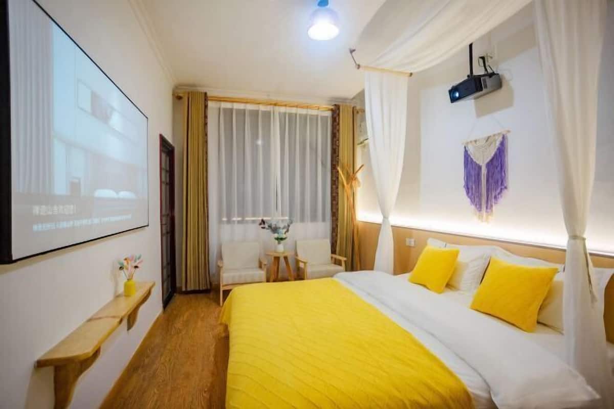 A hotel room with a large bed, a desk, a chair, a projector screen, and a window with curtains.