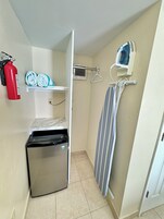Un baño compacto con inodoro, un pequeño cubo de basura, un extintor, un perchero con toallas, una cortina de ducha y un aire acondicionado instalado en la pared.