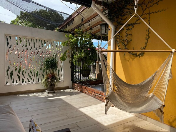 Una hamaca colgada en una pared amarilla, un separador de celosía blanca y plantas en macetas en un patio.
