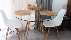 Una mesa de vidrio con dos sillas blancas y un camino de mesa tejido, frente a cortinas translúcidas y un drape gris.