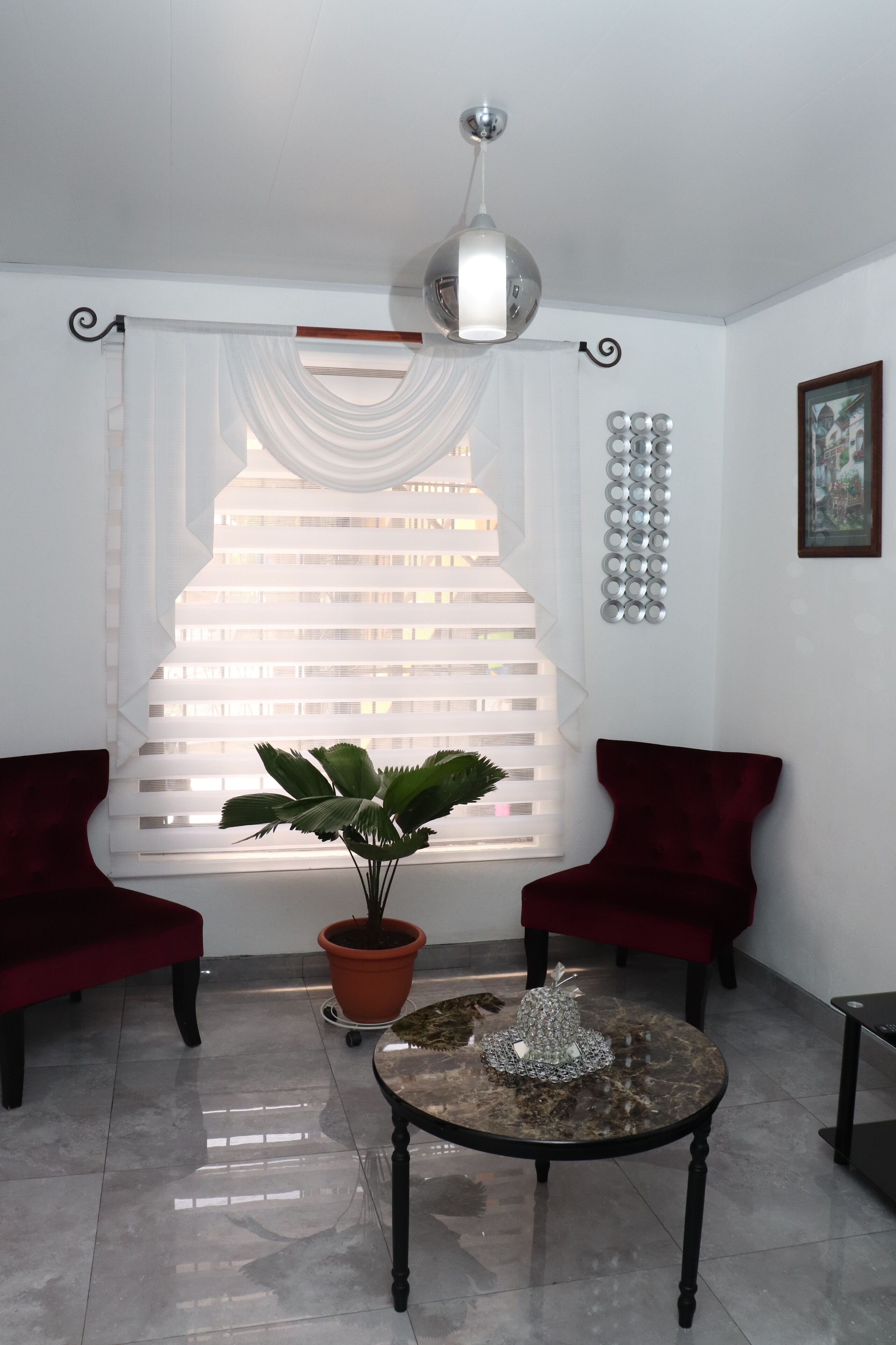 Un salon avec une grande fenêtre, deux fauteuils rouges, une table ornée d’un objet décoratif et une plante en pot.