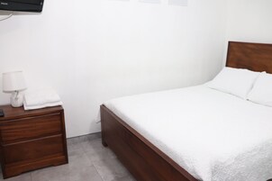 Una cama bien tendida con ropa de cama blanca, una mesita de noche de madera con una lámpara y un televisor montado en la pared.