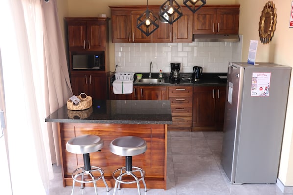 A compact kitchen with wooden cabinets, a black countertop, two bar stools, a stainless steel refrigerator, and a microwave.