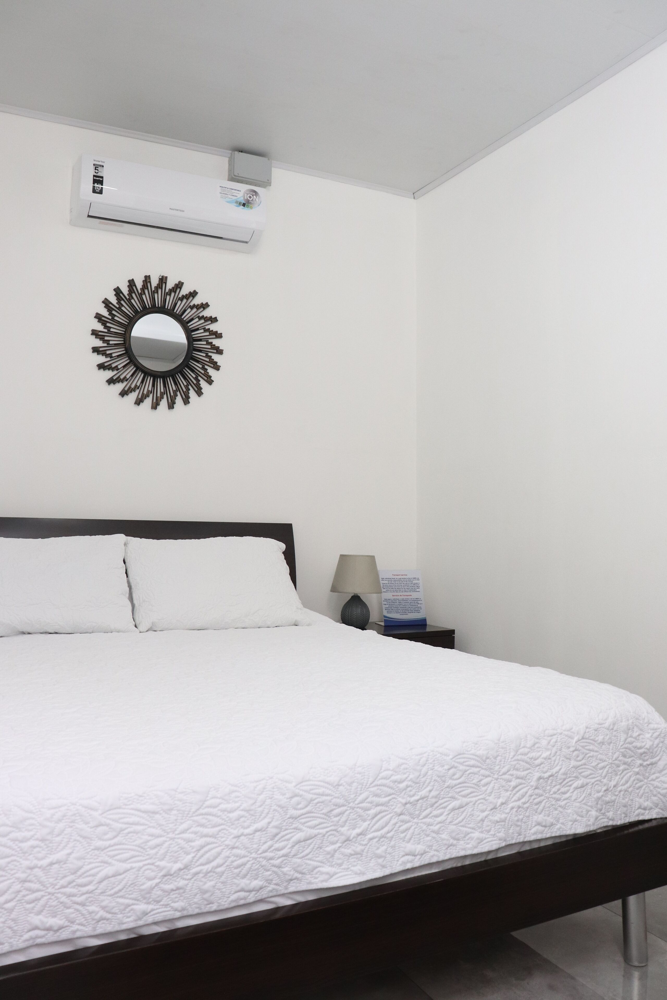 Un lit bien fait, avec du linge de lit blanc, un climatiseur fixé au mur et un miroir décoratif.