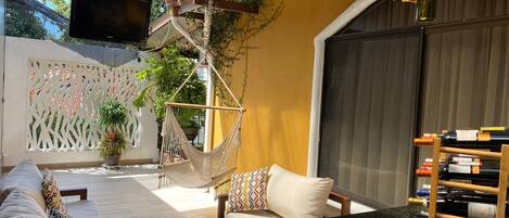 Un patio soleado con una hamaca, mobiliario exterior, un televisor montado en una pérgola de madera y una estantería para vinos.