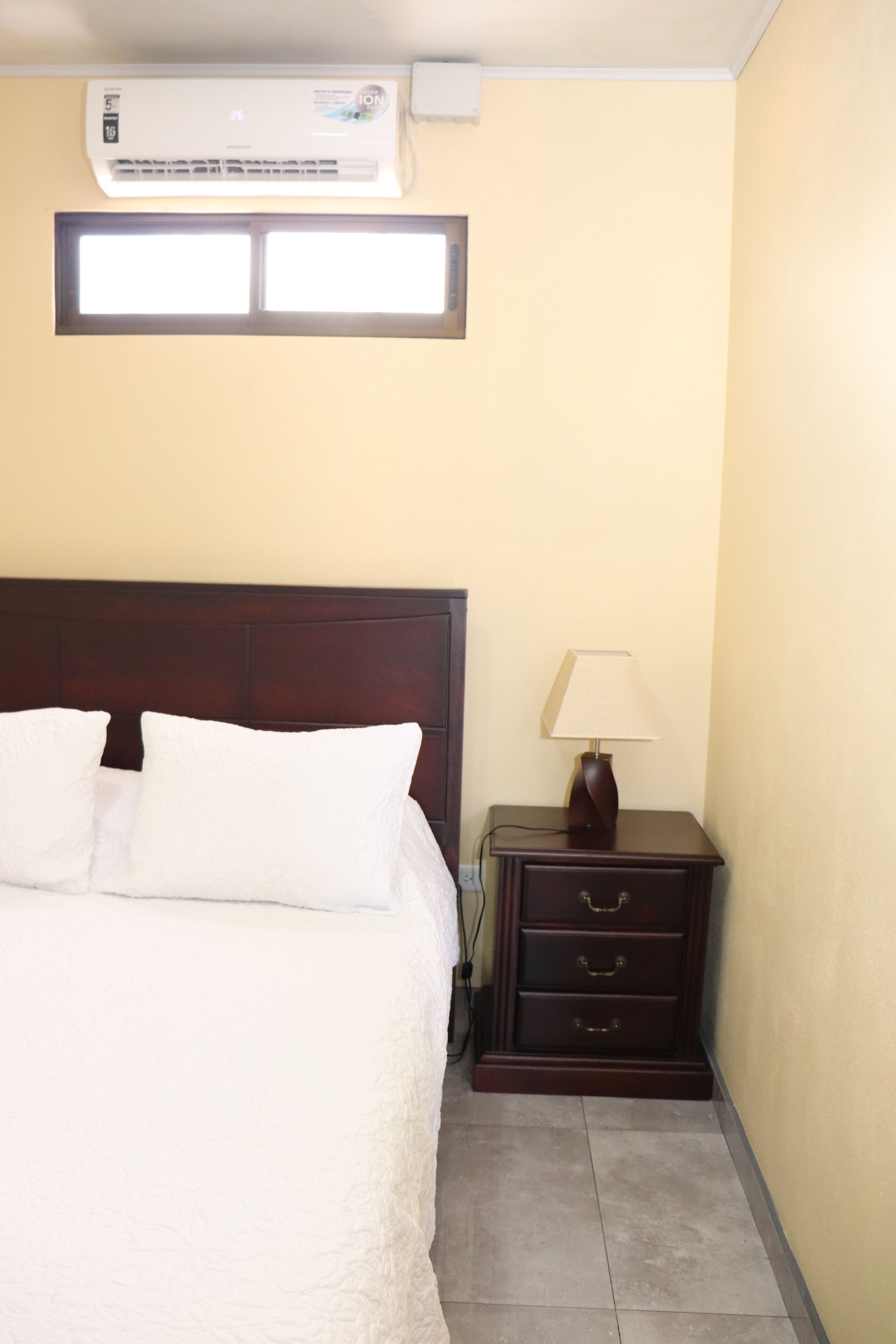 A hotel room with a bed, a nightstand with a lamp, an air conditioner, and a window.