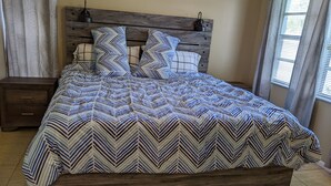 Una cama con colcha estampada y dos almohadas, cabecera de madera, mesita de noche con cajones y una ventana con cortinas.