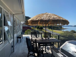 Una terraza de madera con una mesa y sillas, un parasol de paja, y vista a un cuerpo de agua y tierra distante.