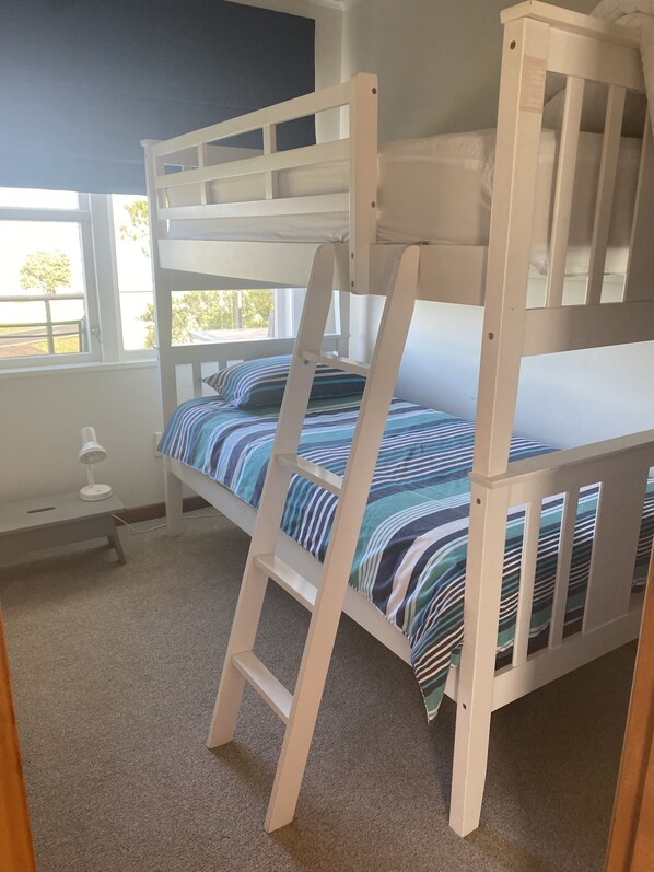 Una cama nido con escalera, ropa de cama a rayas, un escritorio pequeño con lámpara y una ventana con vista a árboles.