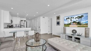 A modern living room with a glass coffee table, a sofa, and a dining area with white furniture. - Spectacular Ocean View Condo in Beachfront Resort (South Padre Island)