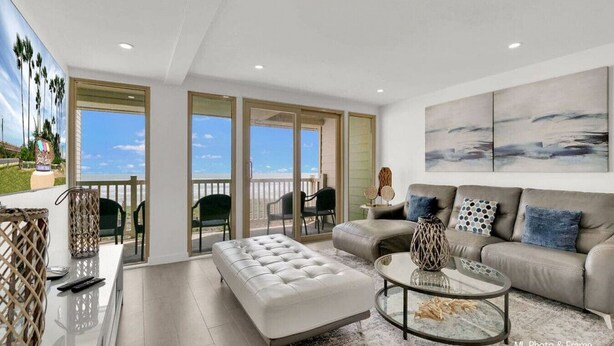 A spacious living room with a large window, a sofa, a coffee table, and a balcony with outdoor seating. - Spectacular Ocean View Condo in Beachfront Resort (South Padre Island)