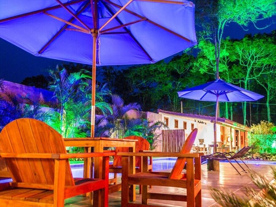 A poolside area with wooden chairs and umbrellas, surrounded by palm trees and a building with a pool.