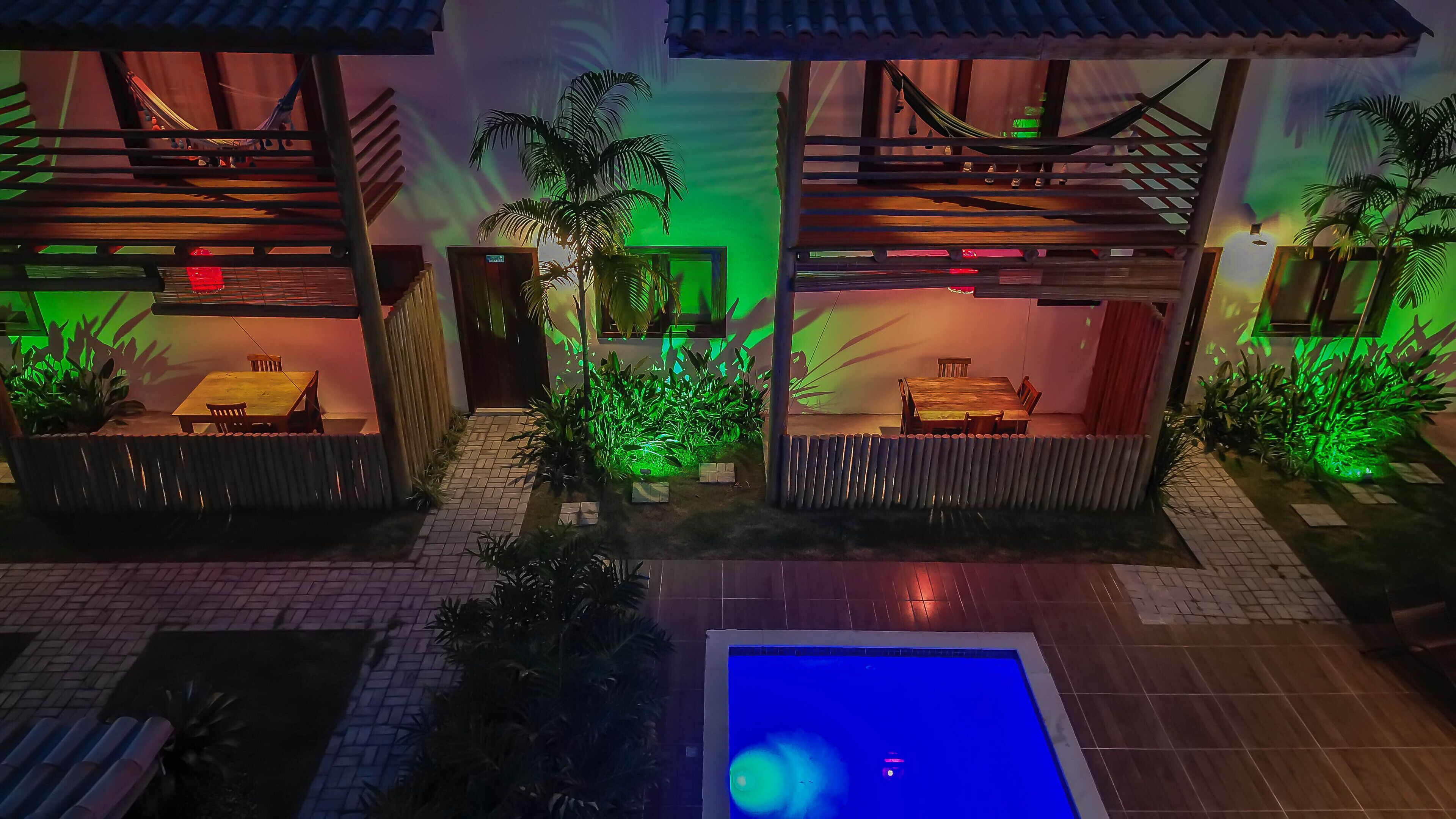 Zona piscina con pavimento piastrellato e piscina. Due strutture in legno con posti a sedere e un piccolo tavolo, circondate da piante e una recinzione.