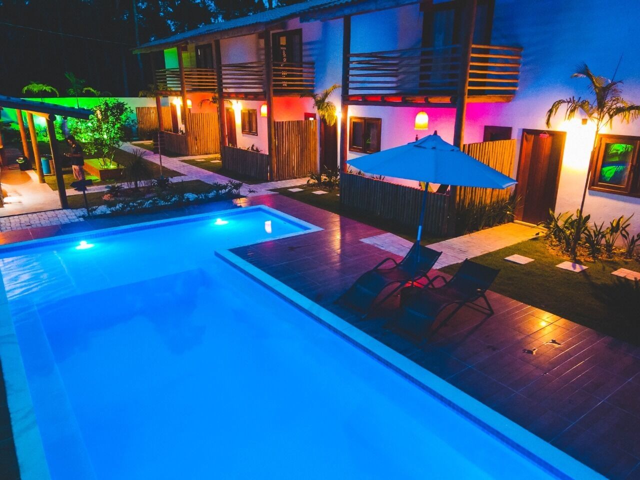 A swimming pool with a wooden deck and lounge chairs, surrounded by a building with outdoor lighting.